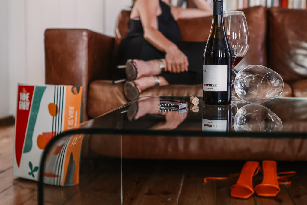 Wine bottle, glasses, and a Wine Fairy gift box styled on a leather couch, an orange heel in the reflection